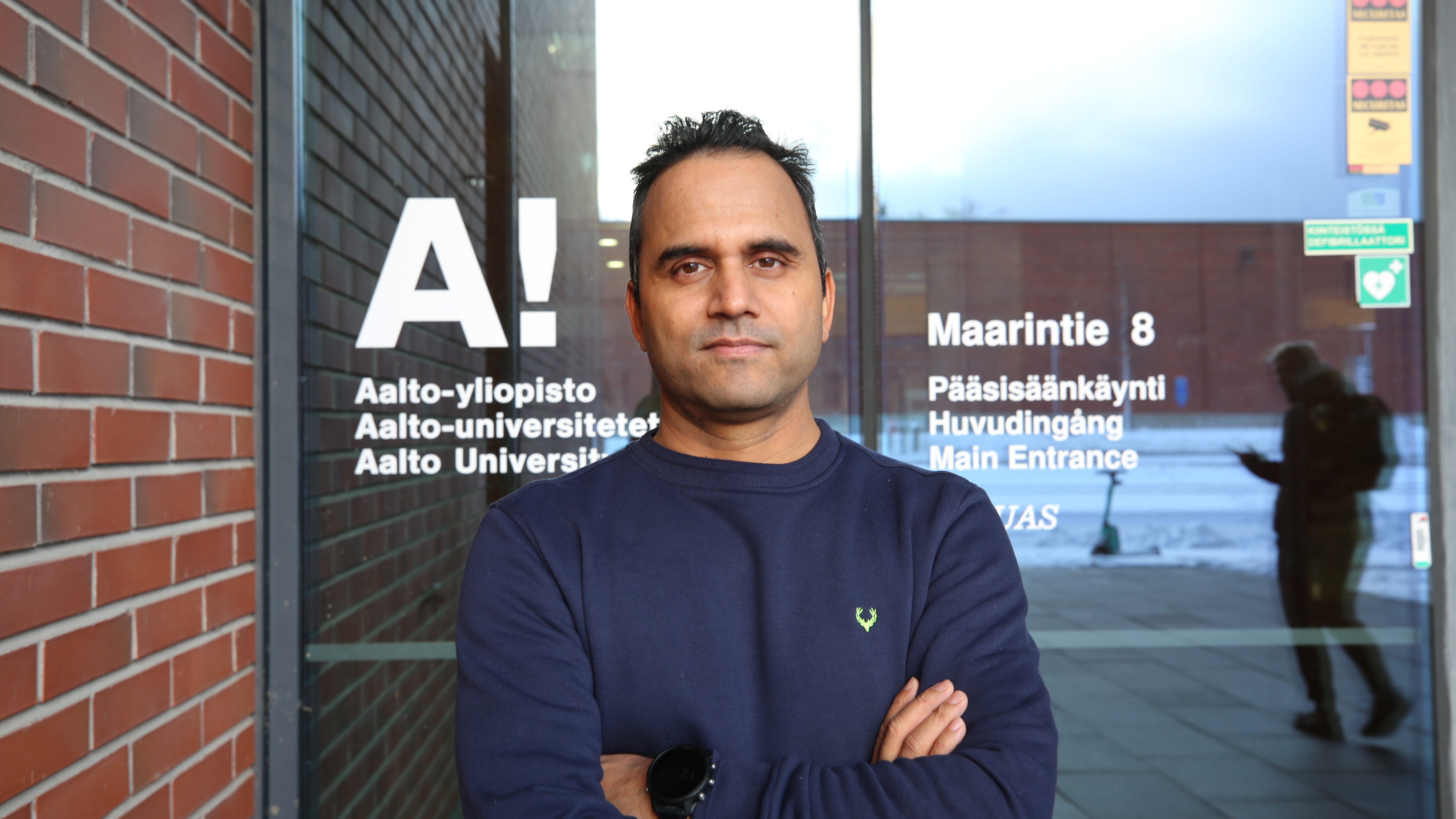 Ristissä käsissä seisova henkilö Aalto-yliopiston sisäänkäynnin edessä. Takana tiiliseinä ja lasiovi tekstillä.