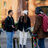 Three people with backpacks are standing by rental bicycles outside a brick building.