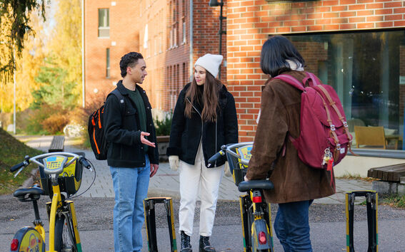 Three people with backpacks are standing by rental bicycles outside a brick building.