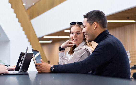 Two people sitting at a table with laptops in a modern building with wooden accents and white walls.