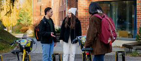 Three people with backpacks are standing by rental bicycles outside a brick building.