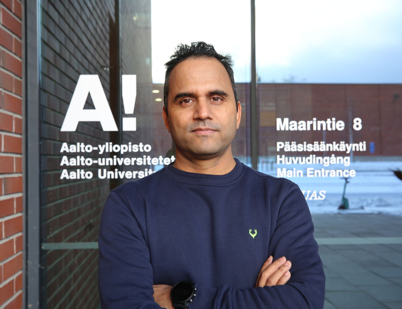Ristissä käsissä seisova henkilö Aalto-yliopiston sisäänkäynnin edessä. Takana tiiliseinä ja lasiovi tekstillä.